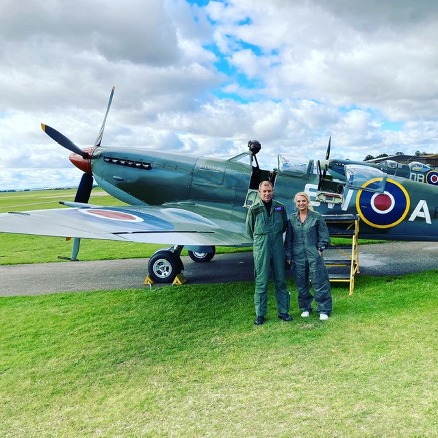 Liv Perry and pilot wear flight suits in-front of a Spitfire.
